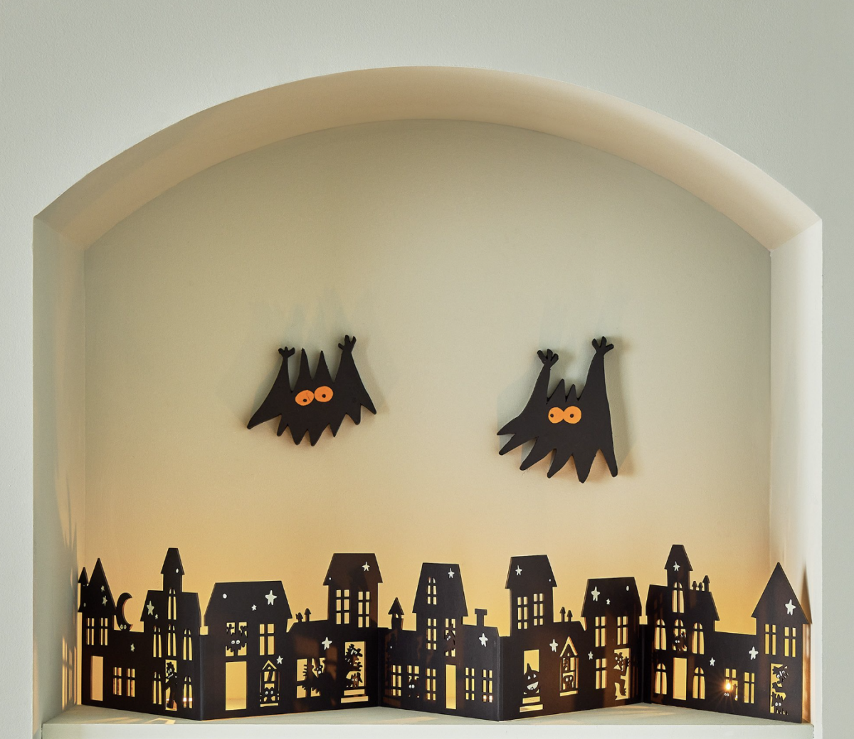 20 Halloween Decor FINDS That Are Actually Chic - 20 Black paper cutouts of bats with orange eyes hang above a row of illuminated black paper houses with Halloween-themed silhouettes, arranged inside a wall alcove—the perfect Halloween decor to celebrate the spooky season.