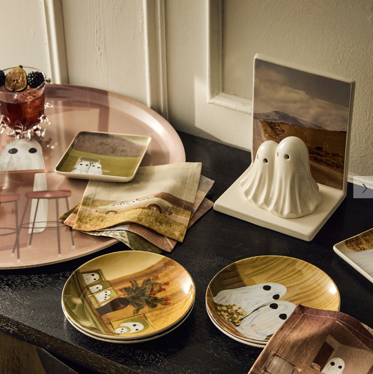 20 Halloween Decor FINDS That Are Actually Chic - 4 A table set with plates, napkins, and trays featuring ghost illustrations, a ghost-shaped salt and pepper shaker, and a glass with fruit, all arranged on a dark surface—perfect for showcasing your spookiest Halloween decor this spooky season.