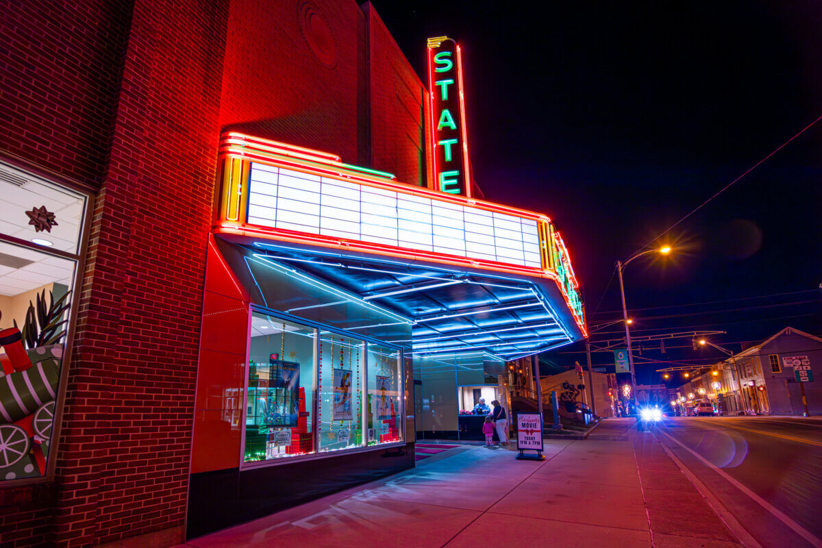 Our Favorite Historic Downtowns in the South to Explore - 9 State Theater with neon lights illuminated at night, marquee sign visible above the sidewalk, people near the entrance, and streetlights lining the road—capturing the lively atmosphere often found in historic downtowns of the South.
