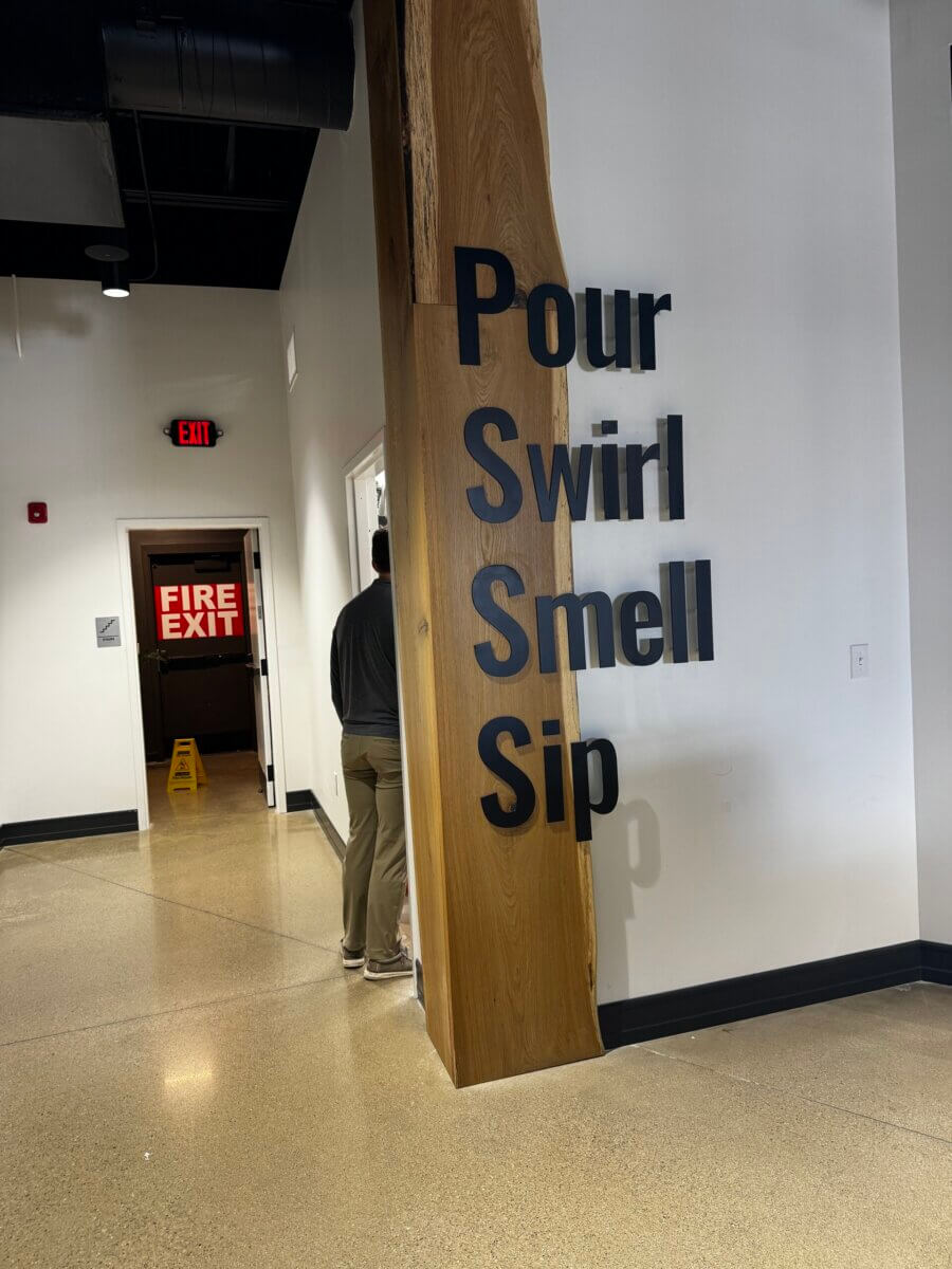 The Commons: Lexington’s Next Big Gathering Space - 4 Large text on a wooden beam reads “Pour, Swirl, Smell, Sip,” welcoming guests into this inviting gathering place. A person stands near a doorway; a fire exit sign and wet floor sign are visible in the background, adding to the everyday charm of this Lexington locale.