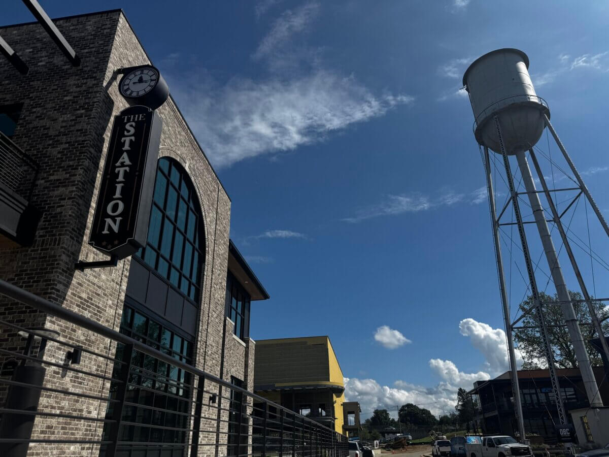 The Commons: Lexington’s Next Big Gathering Space - 5 Brick building with a large clock sign reading “The Station” on the left, a water tower on the right, and a clear blue sky with scattered clouds overhead—this vibrant spot in Lexington is known as The Commons, a popular gathering place for the community.