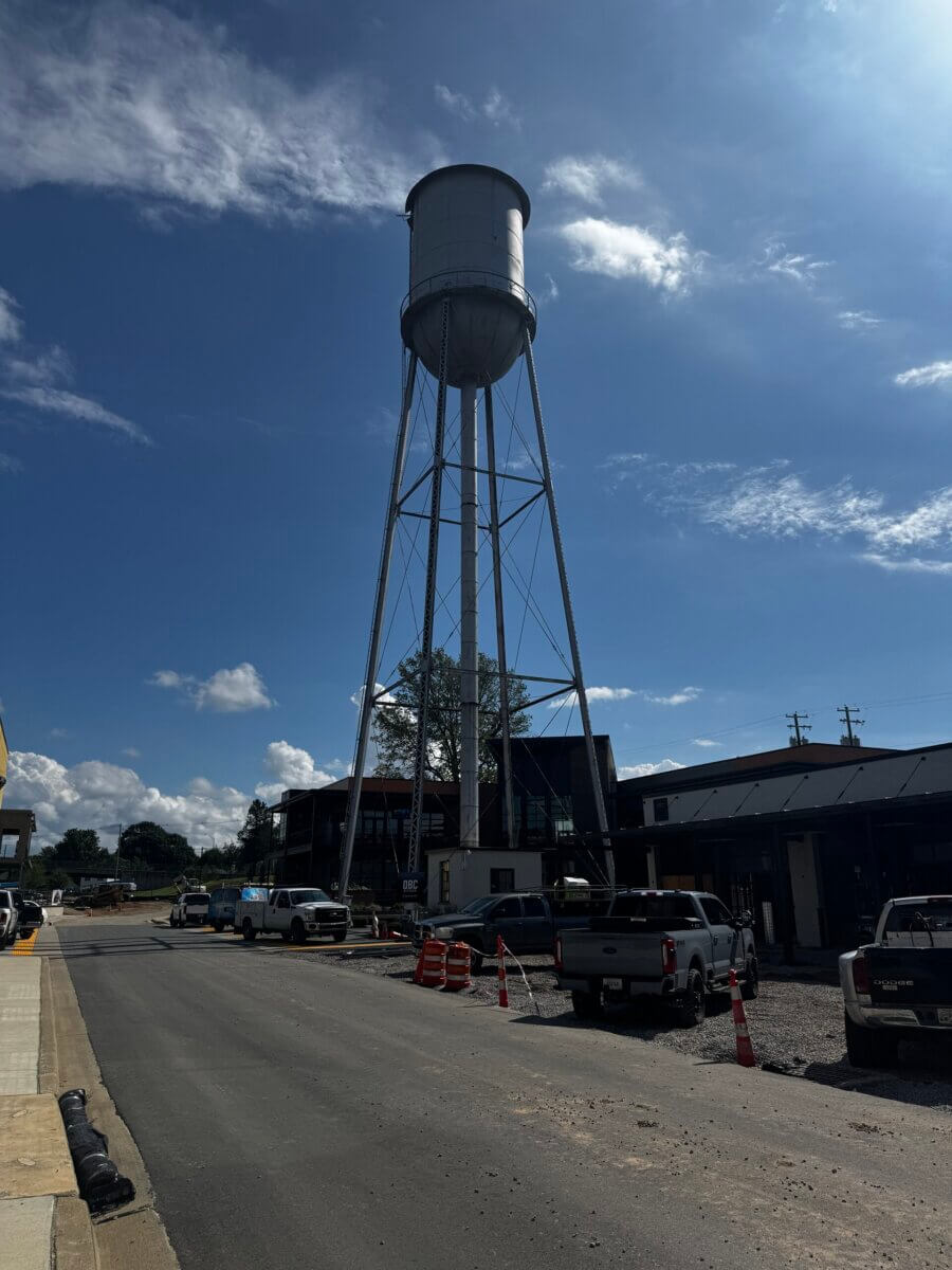 The Commons: Lexington’s Next Big Gathering Space - 3 A tall metal water tower stands over a street with parked vehicles and orange traffic cones on a sunny day, marking the heart of Lexington’s The Commons—a popular gathering place for the community.