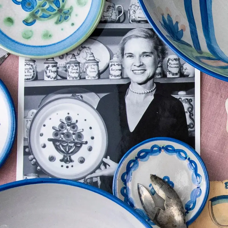 Black-and-white photo of a smiling woman holding a decorated plate, surrounded by colorful ceramic dishes and a metal tool in the foreground.