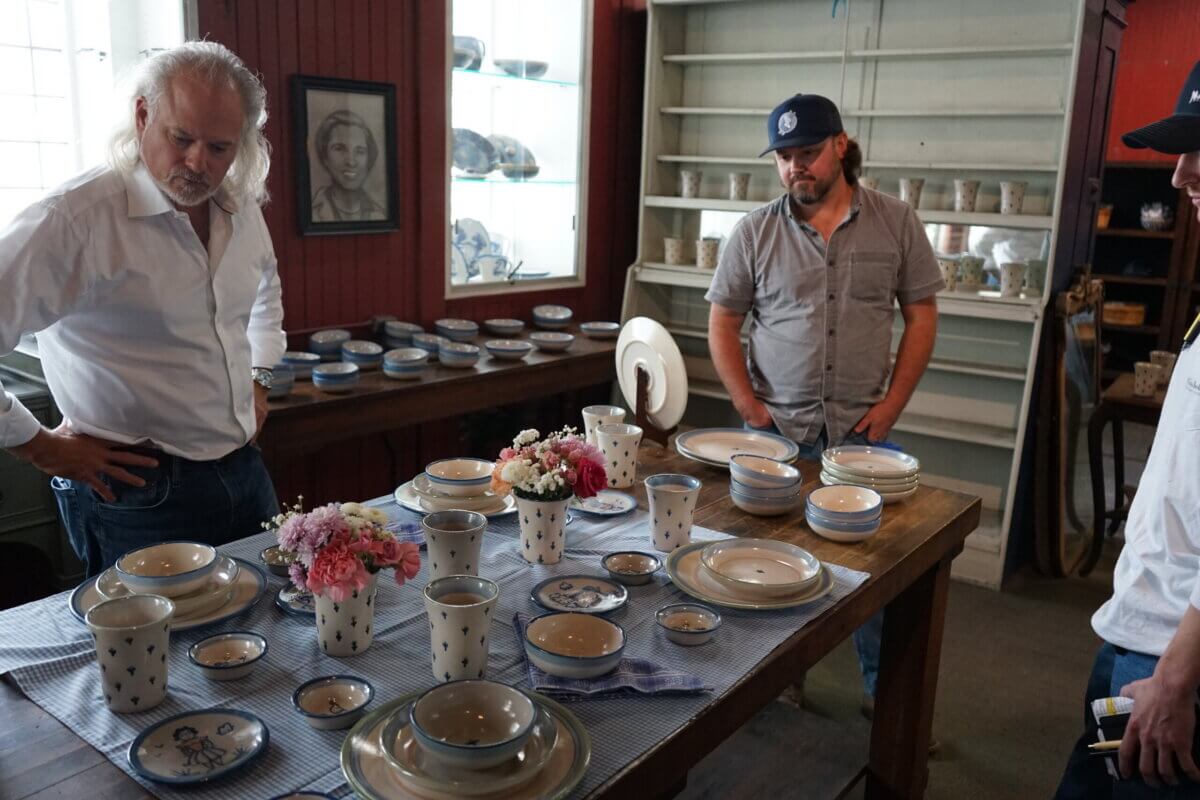 Three men stand around a table set with assorted ceramic dishware and vases of flowers in a room with shelves of pottery and a framed portrait on the wall.