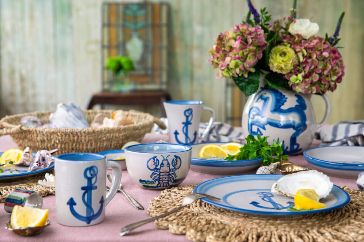 Table set with blue and white anchor-themed dishes, cups, seafood, lemon wedges, and a vase of flowers on a pink cloth with woven placemats.