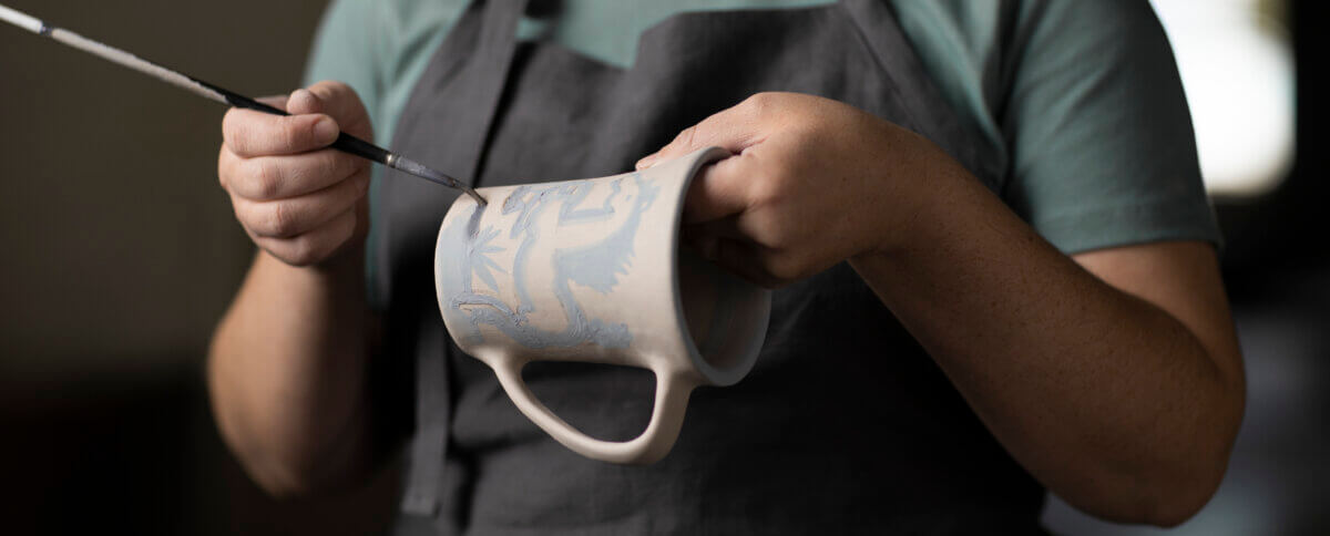 Person wearing an apron holding a ceramic mug and painting a design on it with a thin brush.