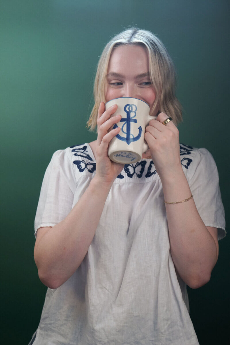A person with blonde hair wearing a white embroidered top holds a mug with a blue anchor design, standing in front of a green background.