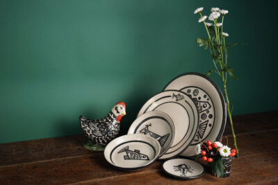 Set of black and white ceramic plates with geometric patterns, a ceramic chicken, and two small vases with flowers, arranged on a wooden table against a green background.