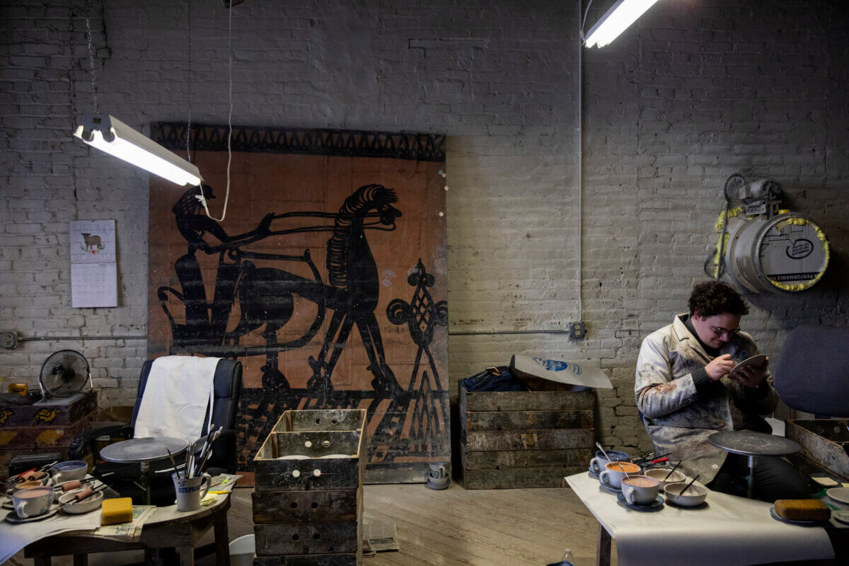 A person sits in an art studio, working on a piece with tools, surrounded by art supplies. A large artwork with horse figures hangs on a brick wall in the background.