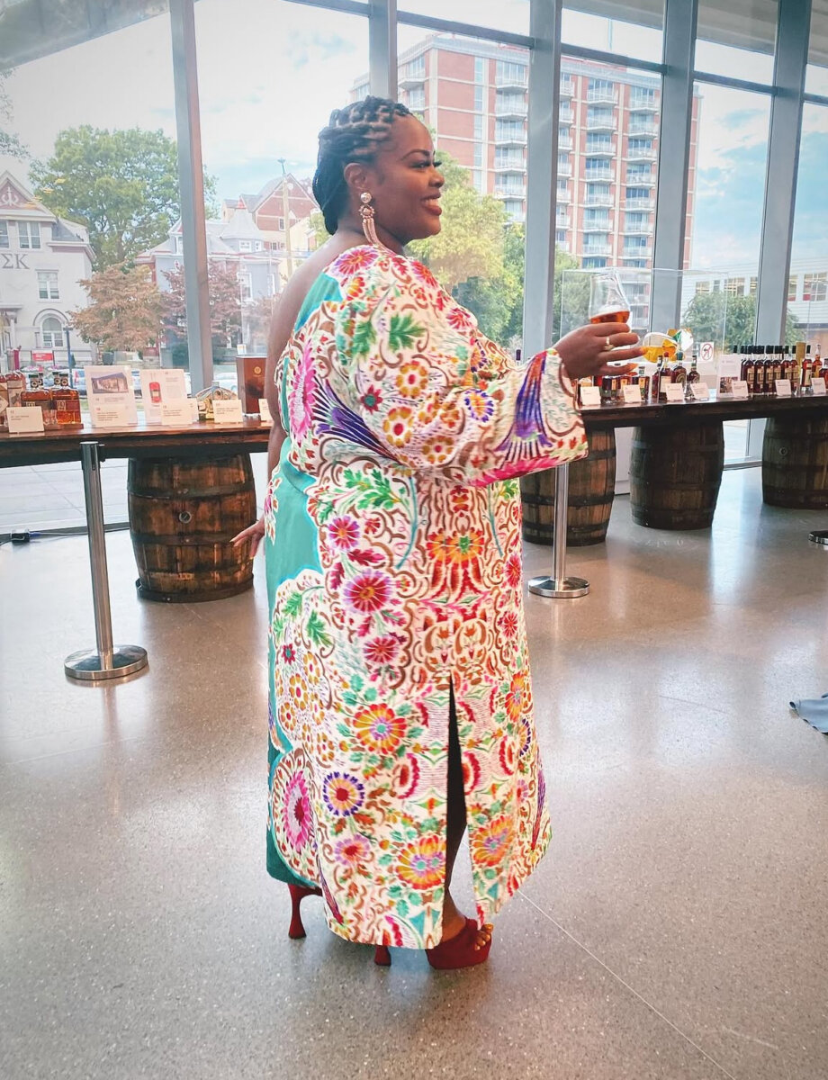 Woman in a colorful floral dress and red heels holds a drink while standing indoors near barrels and a large window with a city view, capturing the vibrant spirit of Louisville’s rich culinary heritage.