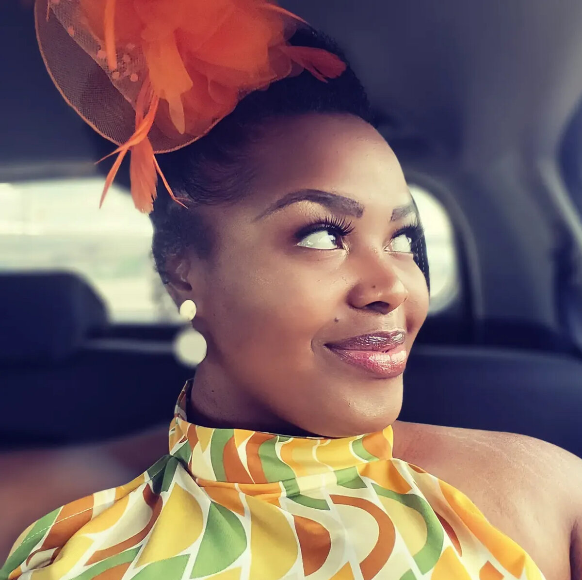 Woman in patterned dress with orange headpiece sits in a car, looking to the side and smiling. She wears pearl earrings and glossy lipstick, embodying the vibrant spirit of Louisville’s rich culinary heritage.