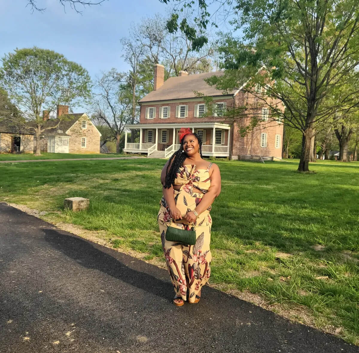 A woman in a floral dress stands on a paved path in Louisville, smiling, with a large brick house and trees in the background under a clear sky.