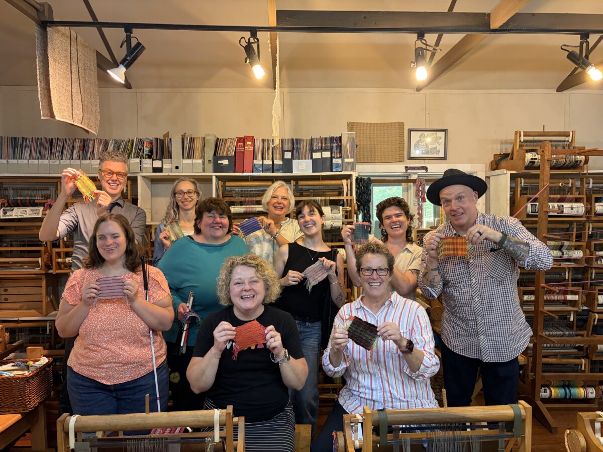 She's Making the Blind Feel Seen Through Louisville’s Newest Museum Experience - 3 Photograph shows nine smiling adults displaying colorful woven samples. The shelves inside a room are filled with yarn, fabric, tools, and wooden looms.