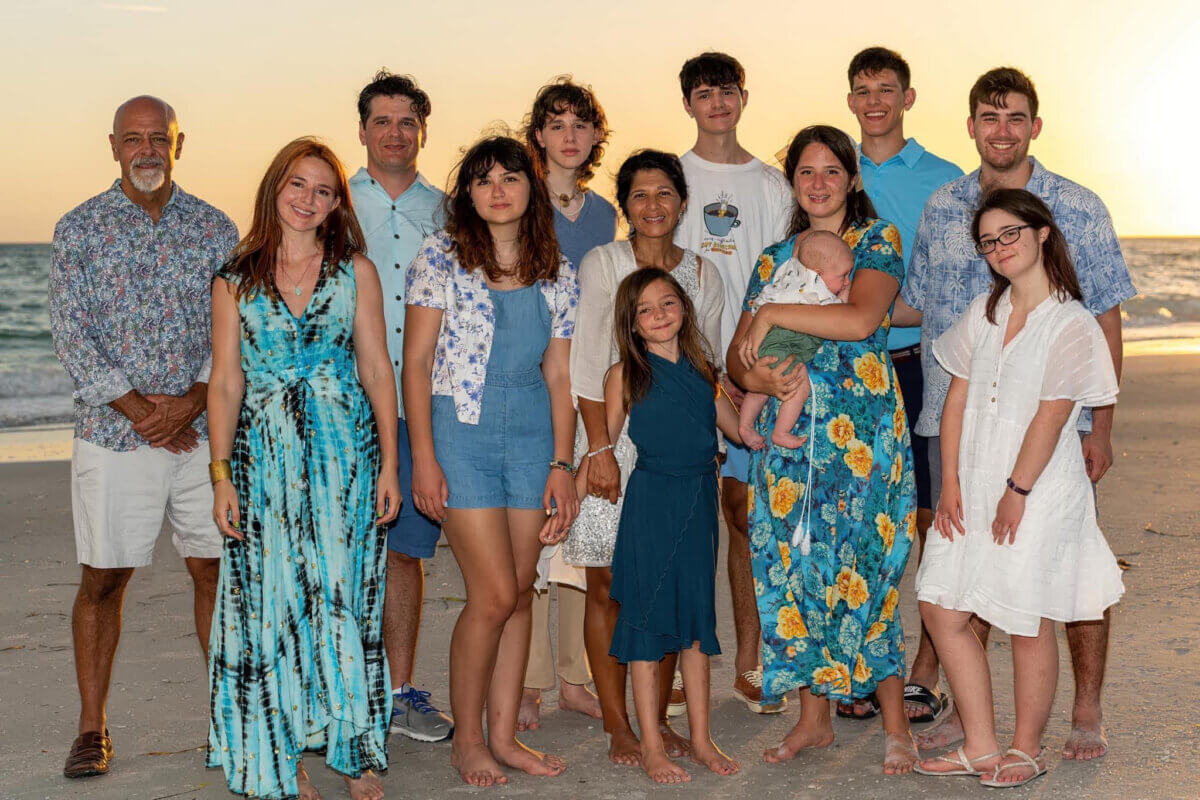 Thirteen people, including a baby, stand barefoot on a beach at sunset, posing for a group photo, with the ocean and sky in the background.