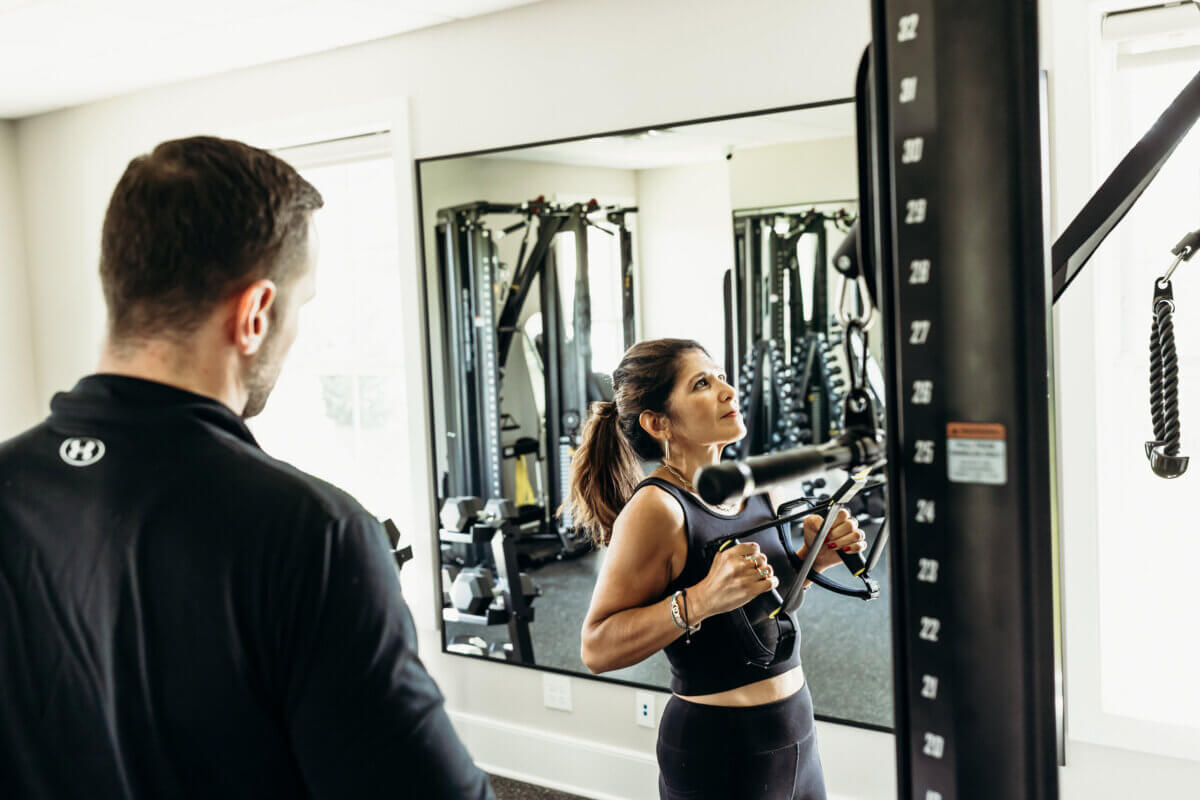 A woman uses a cable machine for strength training while a man observes her in a gym with mirrored walls.