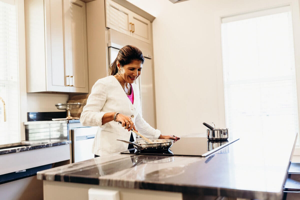 A woman wearing a white blazer cooks at a stove in a modern kitchen, stirring food in a pan while smiling.