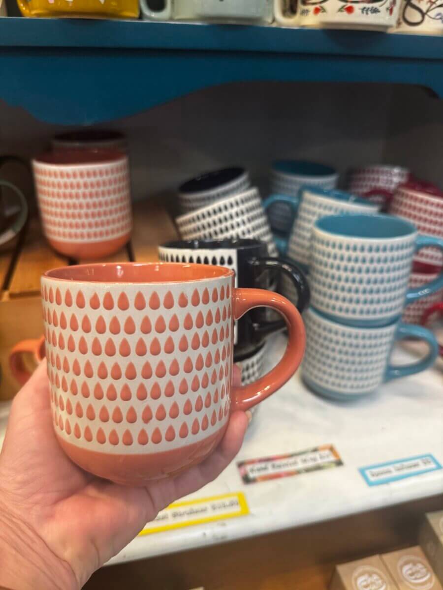 17 Louisville FINDS for October: Fall Fashion, Festive Decor & Game-Day Essentials - 7 A hand holds a coral mug with a white and red teardrop pattern in front of similar patterned mugs stacked on a shelf in a Louisville FINDS store, capturing the cozy spirit of October shopping in Louisville.