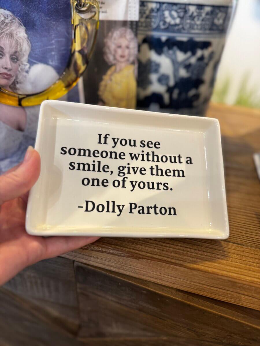 17 Louisville FINDS for October: Fall Fashion, Festive Decor & Game-Day Essentials - 14 A small tray with the quote: “If you see someone without a smile, give them one of yours. -Dolly Parton,” held by a hand on a wooden surface—perfect for cozy finds this October.
