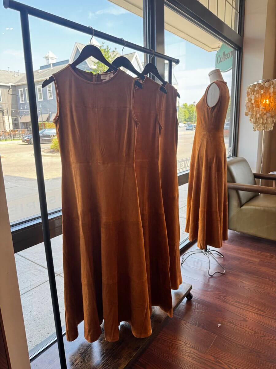 17 Louisville FINDS for October: Fall Fashion, Festive Decor & Game-Day Essentials - 2 Three brown sleeveless dresses hang on a clothing rack near a window, with a matching dress displayed on a mannequin beside them in the boutique—an inviting showcase of autumn-ready FINDS perfect for October in Louisville.