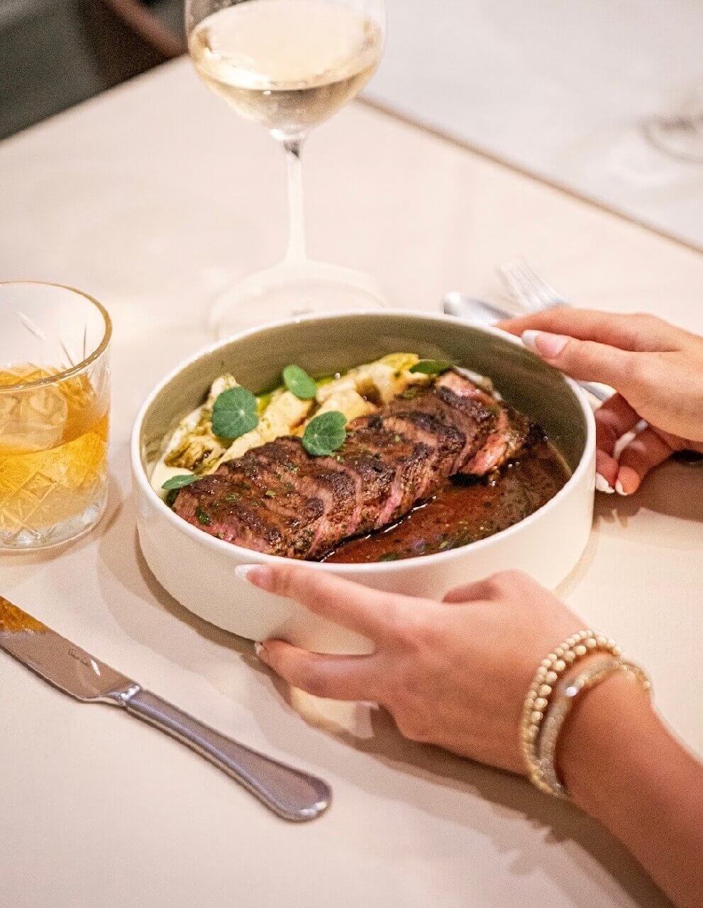 A person holds a bowl with sliced steak and vegetables next to a glass of white wine and a cocktail on a table set with a knife and fork, showcasing the fresh flavors offered by new menus at Louisville restaurants.