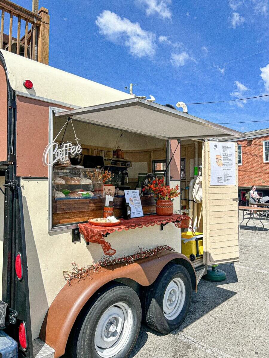 A small coffee trailer with a serving window open, menu displayed, baked goods visible, and flowers on the counter, set in a sunny parking lot—much like the charming new restaurants adding fresh flavor to Louisville’s dining scene with creative new menus.