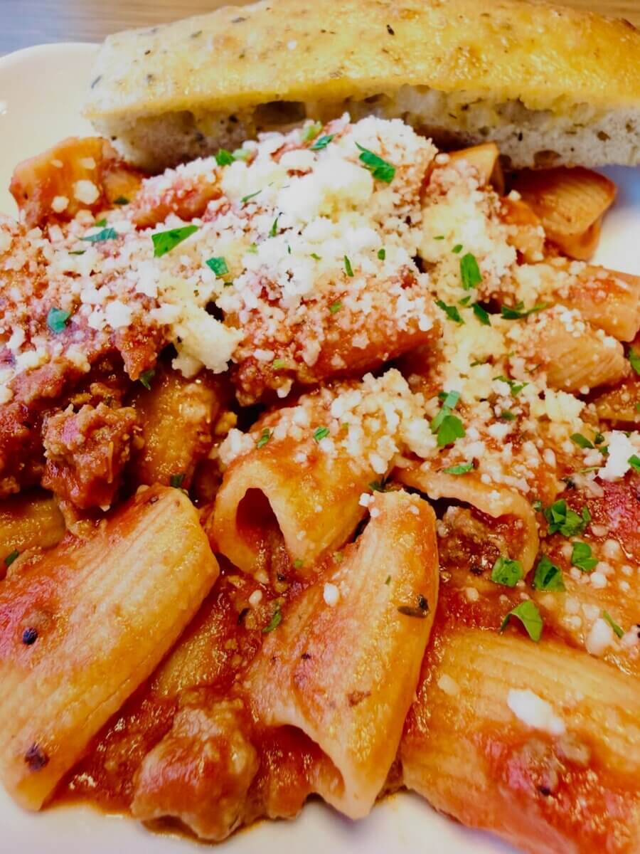 Pasta with tomato sauce and ground meat, topped with grated cheese and herbs, served with a slice of focaccia bread on a white plate—a delicious option often featured on new menus at Louisville restaurants.