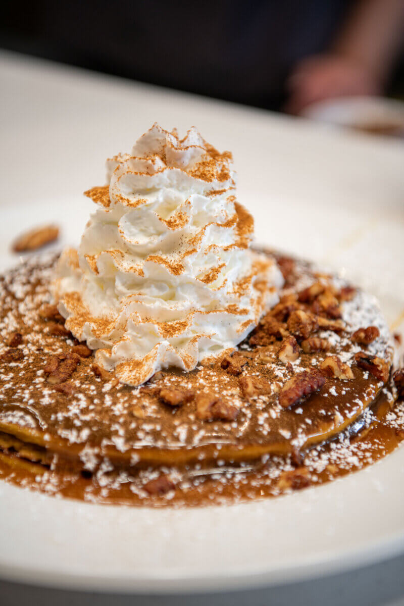 A stack of pancakes topped with whipped cream, powdered sugar, chopped nuts, and syrup on a white plate—perfect for discovering the fresh flavors popping up on new menus at Louisville restaurants.