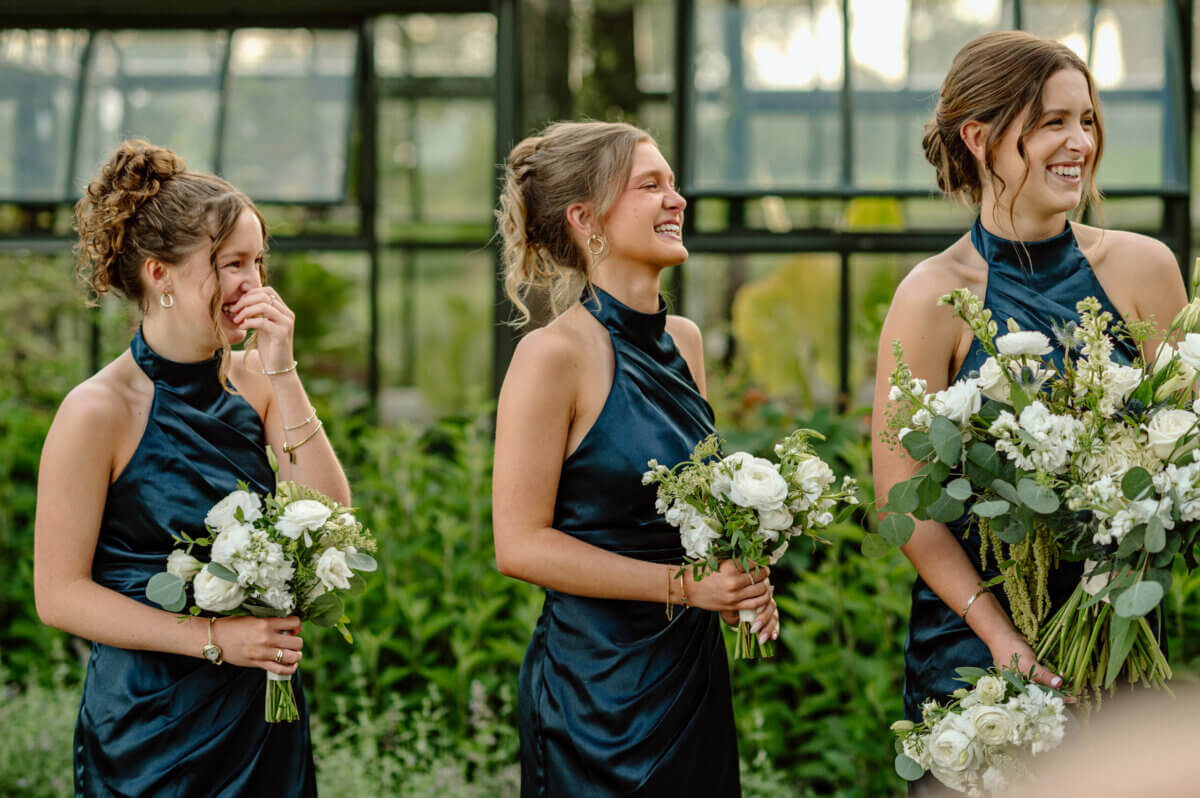 18 Guests + One Gorgeous Louisville Greenhouse Wedding - 5 Three women in matching navy dresses hold white floral bouquets and stand outdoors, smiling and laughing, with greenery and a glass structure in the background—capturing the joyful spirit of a Louisville couple who chose meaning over tradition for their intimate micro-wedding.