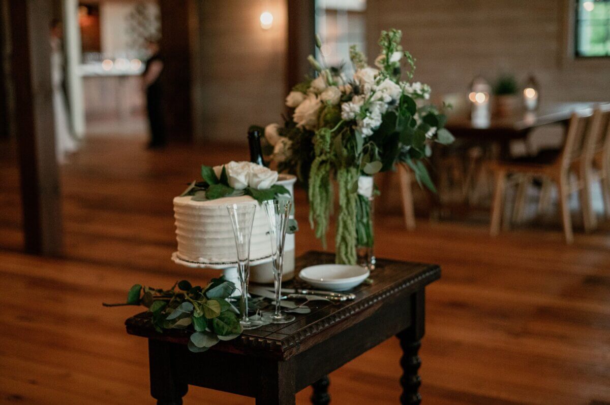 18 Guests + One Gorgeous Louisville Greenhouse Wedding - 12 A small table holds a white frosted cake with flowers, two empty champagne flutes, a bouquet, and plates in a warmly lit wooden room—perfect for a Louisville couple embracing meaning over tradition with their intimate micro-wedding.