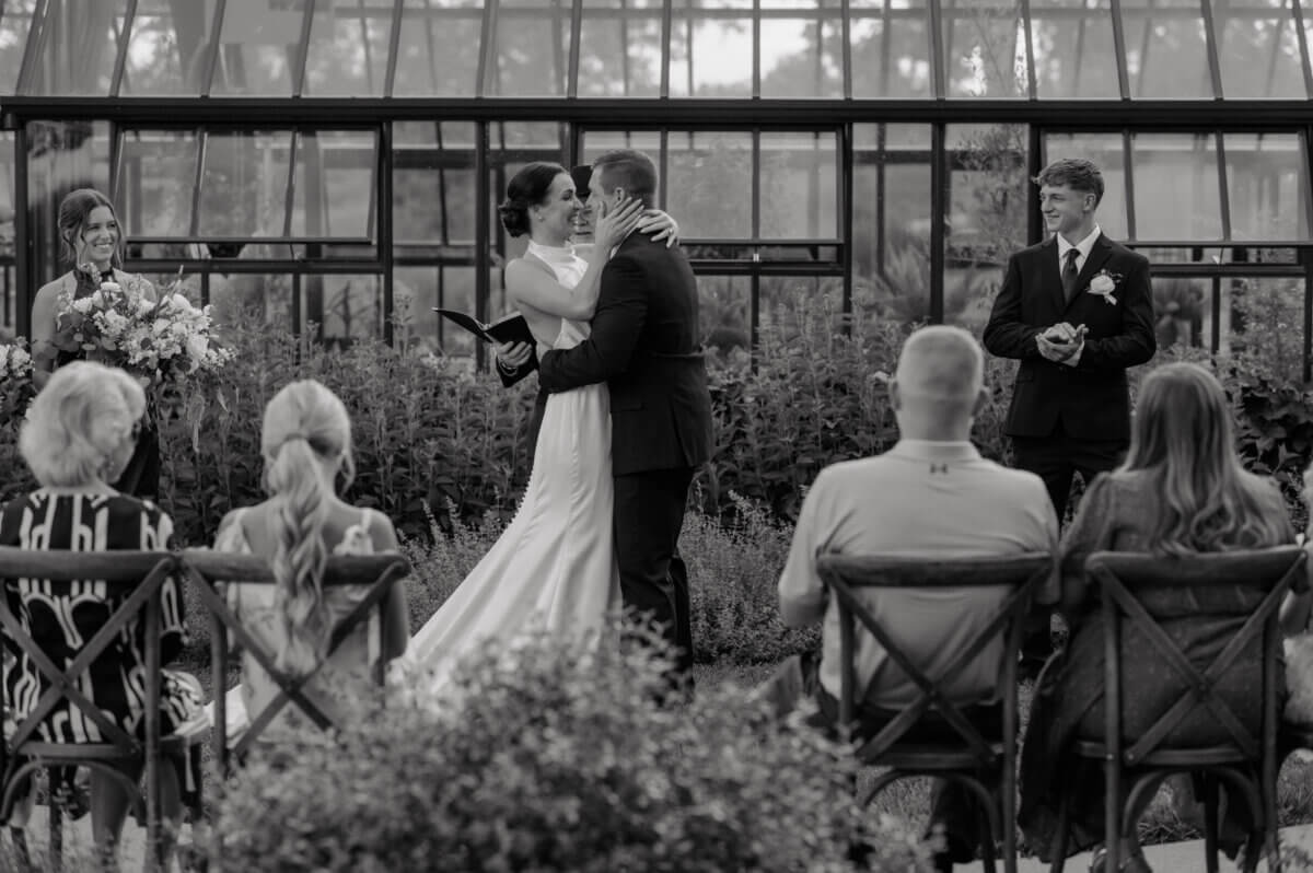 18 Guests + One Gorgeous Louisville Greenhouse Wedding - 4 A Louisville couple shares a kiss during their outdoor micro-wedding ceremony as guests watch, with the bridesmaid and groomsman standing nearby—choosing meaning over tradition on their special day.