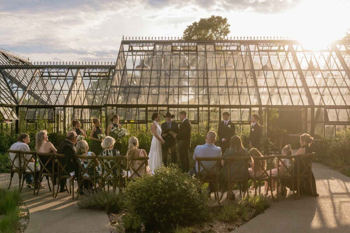 18 Guests + One Gorgeous Louisville Greenhouse Wedding - 9 A small wedding ceremony takes place outdoors in front of glass greenhouses at sunset, with guests seated and the wedding party standing—a meaningful micro-wedding for a Louisville couple who chose meaning over tradition.