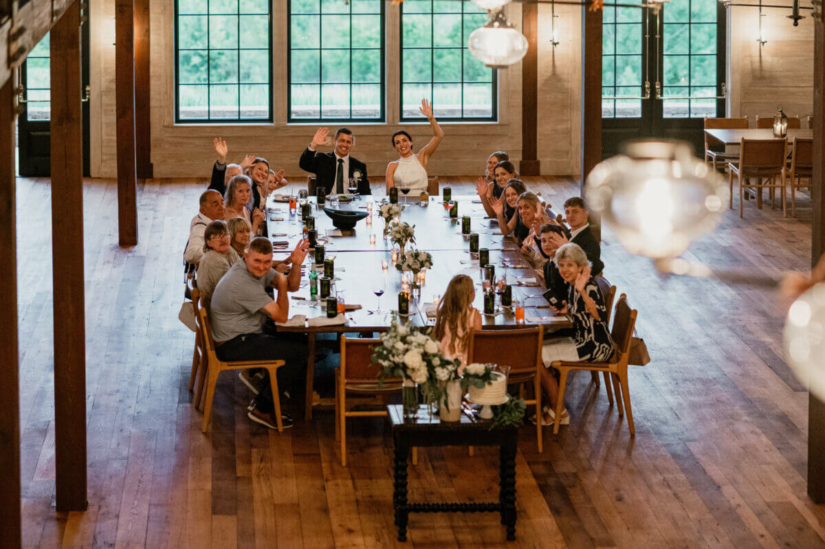 18 Guests + One Gorgeous Louisville Greenhouse Wedding - 14 A group of people sits around a long dining table in a spacious room with wooden floors, waving at the camera during a meaningful wedding celebration.