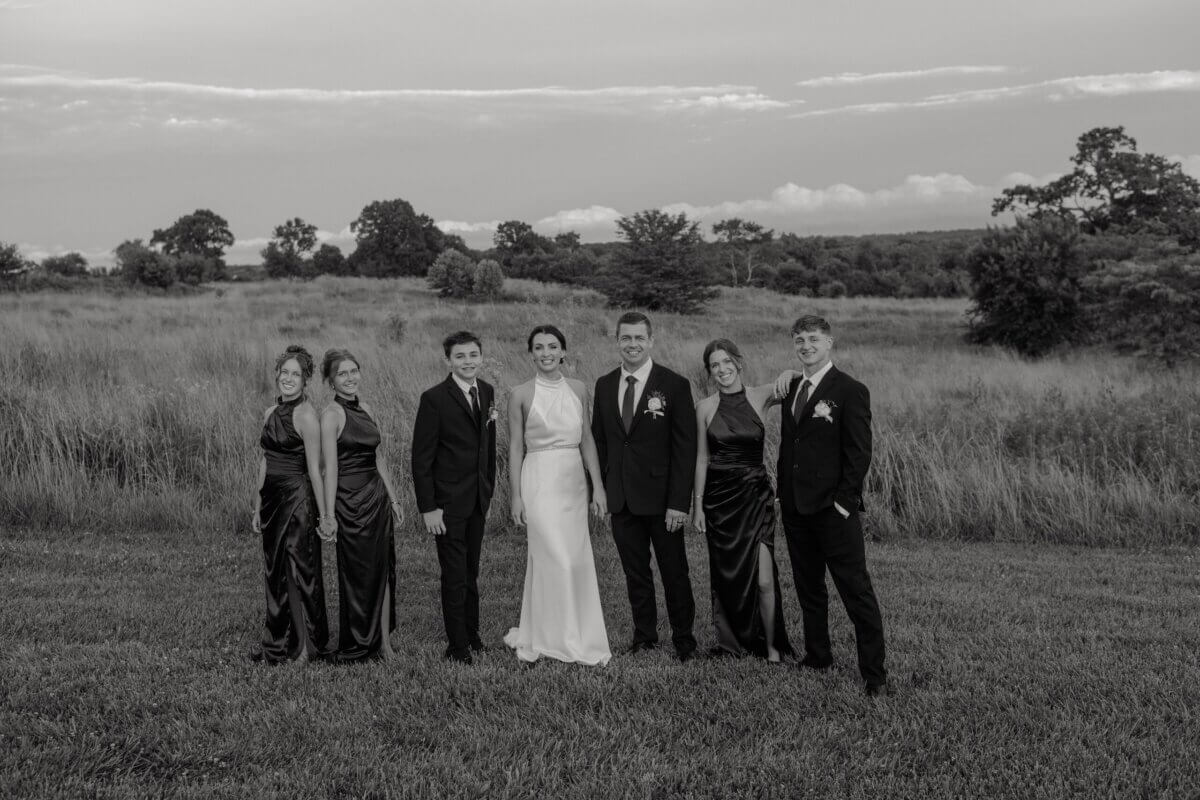 18 Guests + One Gorgeous Louisville Greenhouse Wedding - 3 Six people dressed formally stand in a grassy field, with trees and a cloudy sky in the background; five in dark attire, one in a white dress.