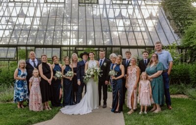 A wedding group photo shows a Louisville couple with family and friends standing in front of a large glass greenhouse, all dressed in formal attire, with greenery in the background—a meaningful moment that beautifully captures their choice of meaning over tradition.