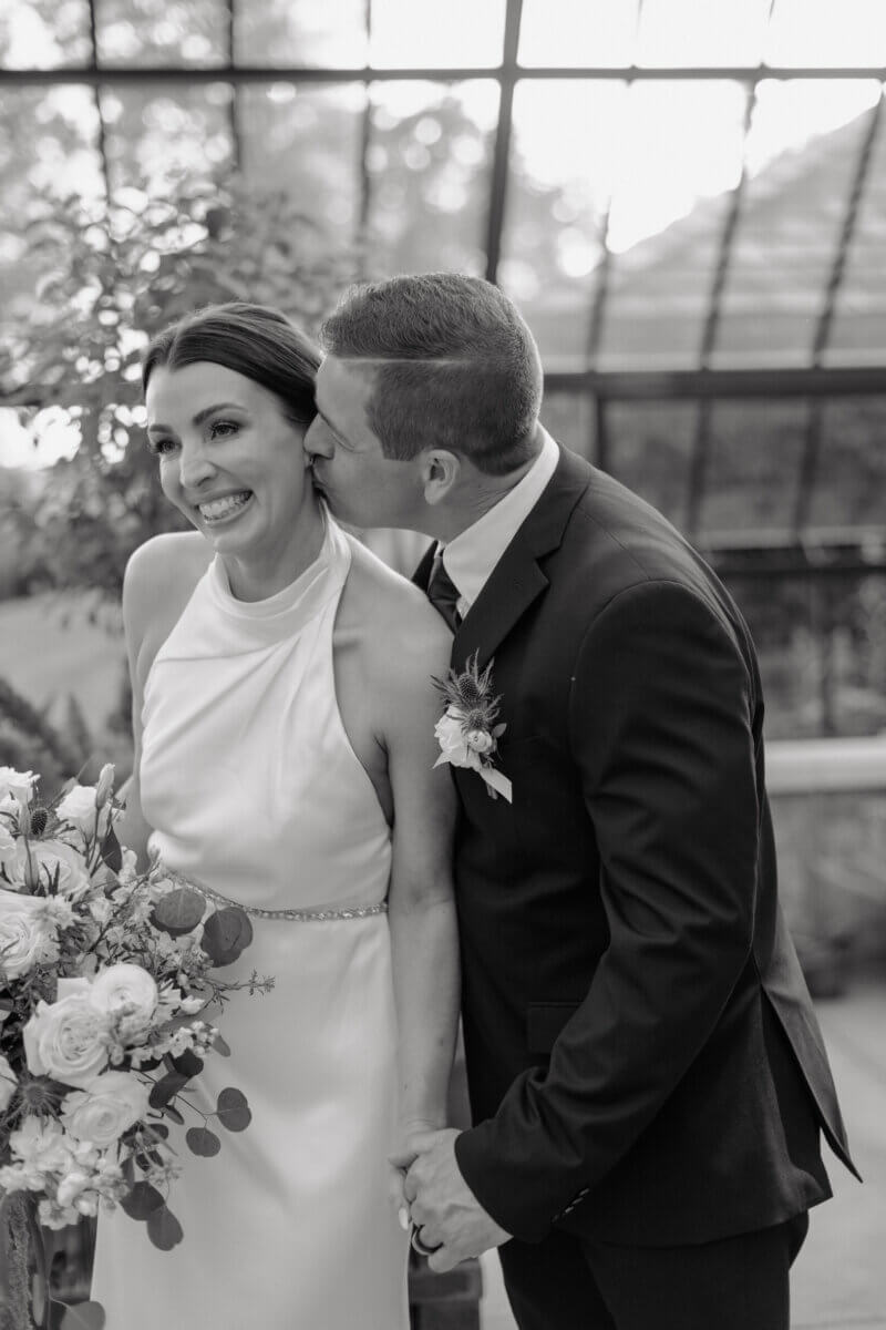 18 Guests + One Gorgeous Louisville Greenhouse Wedding - 10 A Louisville couple embraces meaning over tradition as a groom in a suit kisses his smiling bride on the temple while they hold hands; she holds a bouquet of flowers, capturing the intimacy of their micro-wedding.