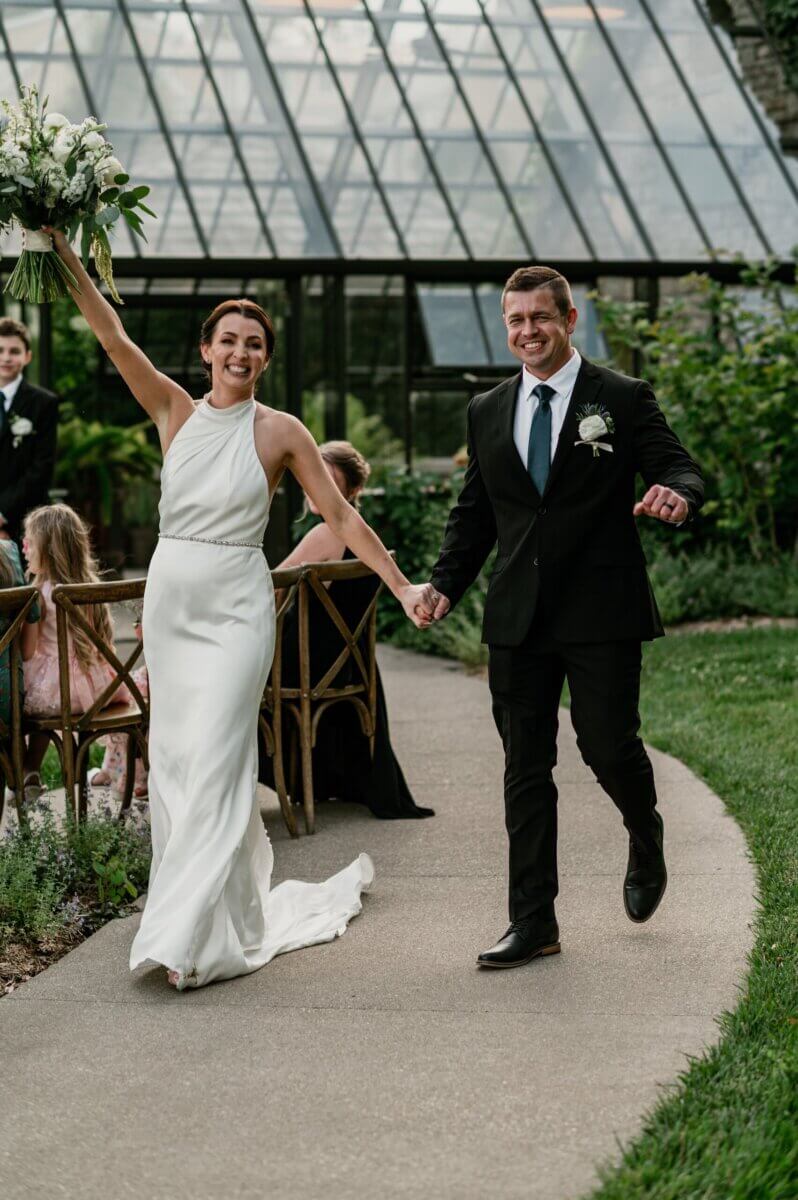 18 Guests + One Gorgeous Louisville Greenhouse Wedding - 2 Bride in a white dress and groom in a black suit walk hand in hand outside, smiling and celebrating their meaningful wedding, with greenery and a glass house in the background.