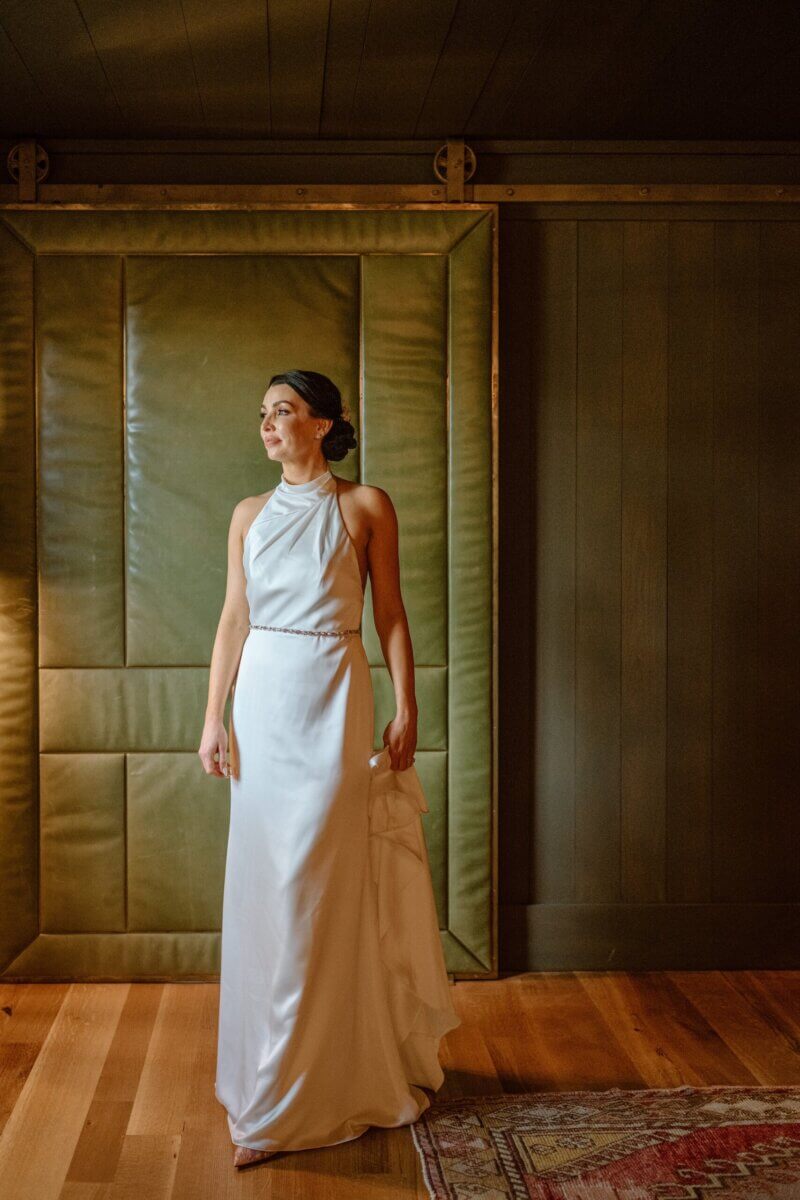 18 Guests + One Gorgeous Louisville Greenhouse Wedding - 6 A woman in a white halter-neck gown stands barefoot on a wooden floor, holding part of her dress, with padded green panels and dark wood walls in the background—a serene moment that captures the spirit of meaning over tradition, perfect for an intimate micro-wedding.