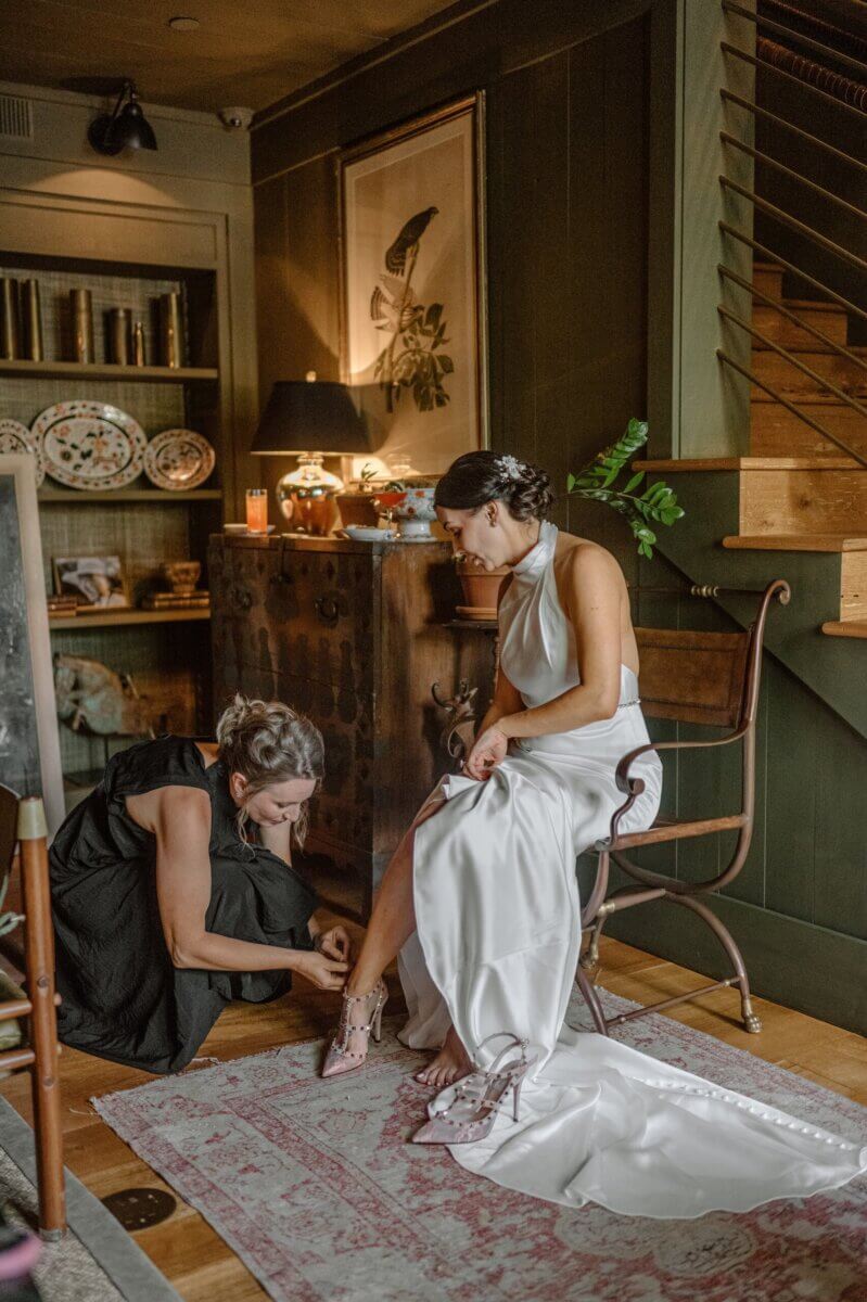 18 Guests + One Gorgeous Louisville Greenhouse Wedding - 7 A Louisville couple celebrates their micro-wedding as a woman in a white wedding dress sits on a chair, while another woman kneels to help fasten her shoes in a warmly lit room with dark walls and wooden furniture.