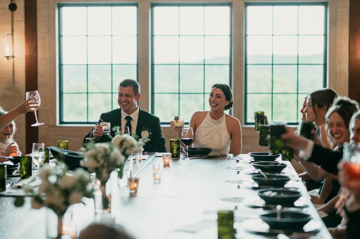 18 Guests + One Gorgeous Louisville Greenhouse Wedding - 11 A group of people in formal attire sit around a long table, raising glasses in a toast, as a Louisville couple celebrates their micro-wedding with meaning over tradition, framed by large windows in the background.