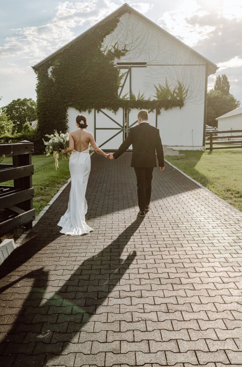 18 Guests + One Gorgeous Louisville Greenhouse Wedding - 15 A bride and groom walk hand-in-hand down a brick path toward a white barn covered in ivy, sunlight casting long shadows behind them—a perfect moment for their meaningful wedding in Louisville, embracing the beauty of an intimate micro-wedding.