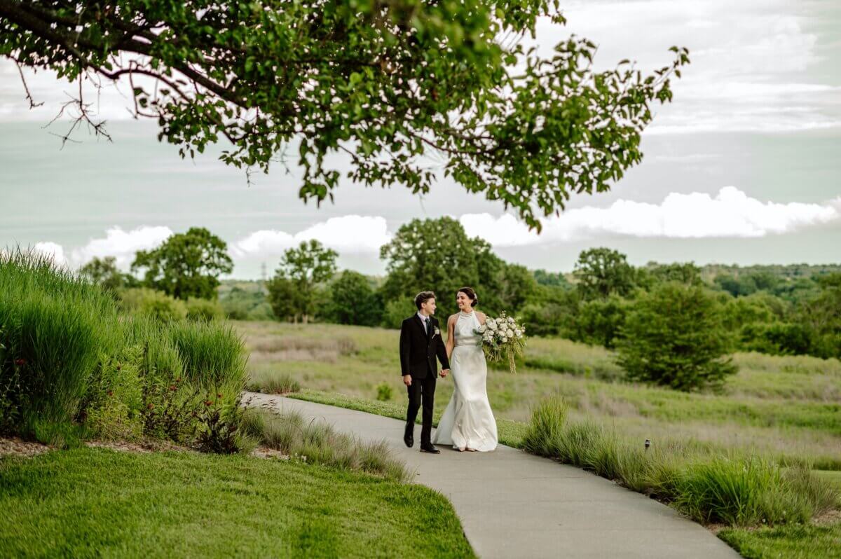 18 Guests + One Gorgeous Louisville Greenhouse Wedding - 8 A Louisville couple walk hand in hand on a paved path through a green field, with trees and a cloudy sky in the background, embracing meaning over tradition as they celebrate their micro-wedding.