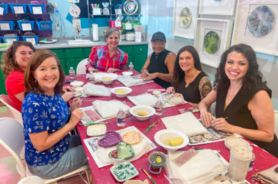 Six women sit around a table with pottery supplies, smiling at the camera in a brightly lit art studio as they connect and create together—seizing the clay with laughter and creativity.