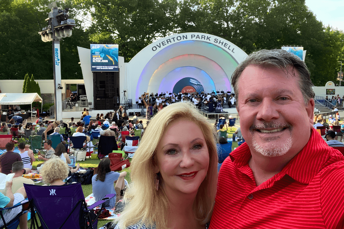 A man and woman pose for a selfie at an outdoor concert in front of the Overton Park Shell stage, surrounded by a crowd seated on the lawn.