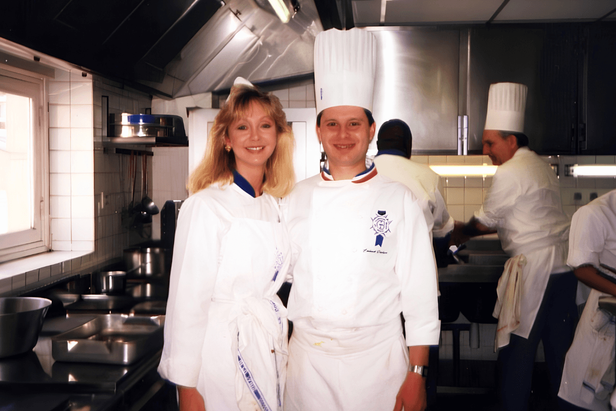 Two chefs in white uniforms and hats stand smiling in a professional kitchen, with other chefs working in the background near stainless steel counters and appliances.