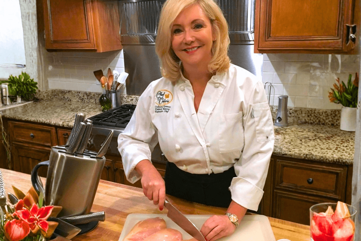 A woman in a chef’s coat slices raw chicken breasts on a cutting board in a home kitchen with wooden cabinets.