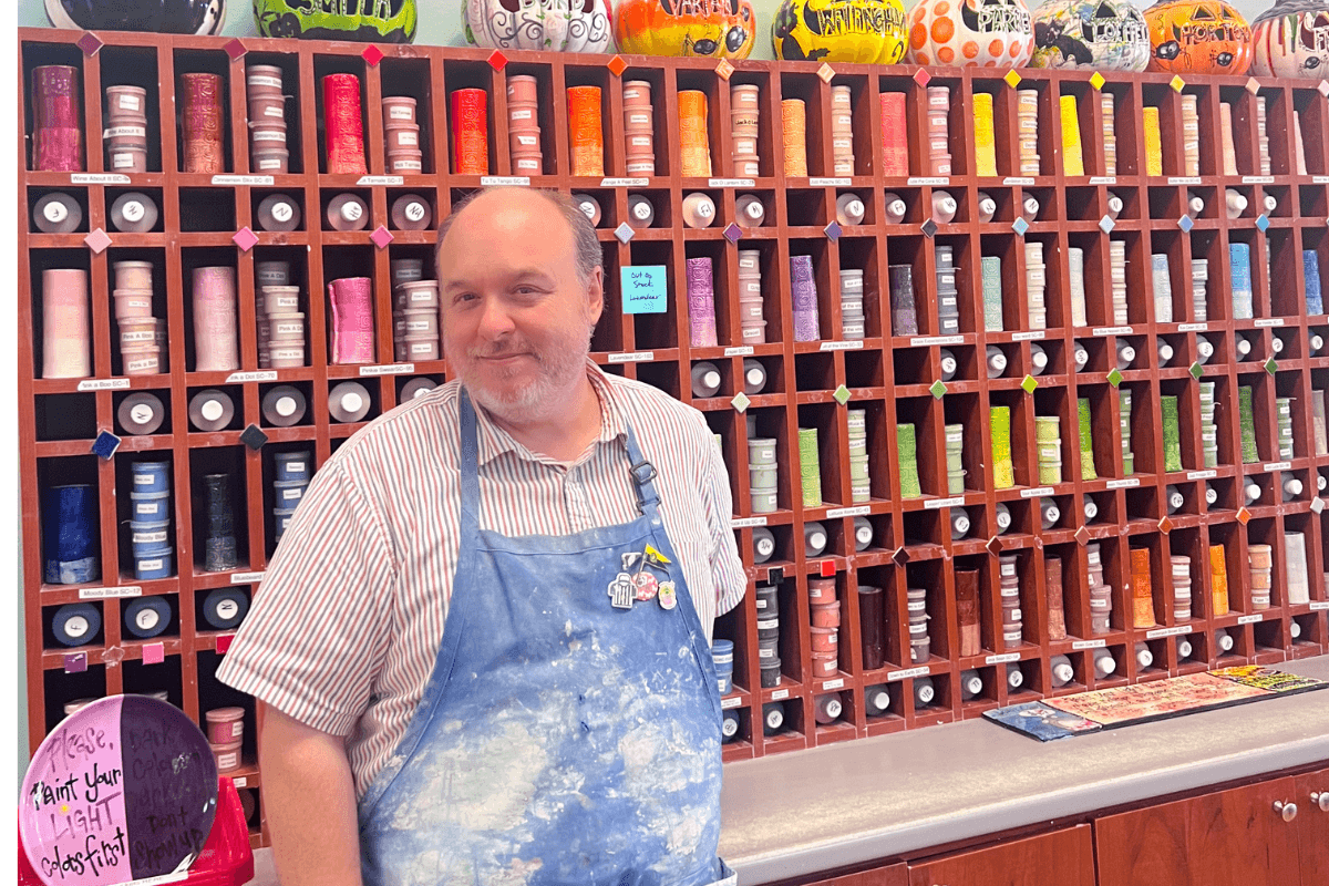 Create, Connect & Get a Little Messy at This Memphis Pottery Studio - 4 A man wearing a paint-stained apron stands in front of shelves filled with colorful ceramic glaze bottles, with painted pottery displayed on the top shelf.