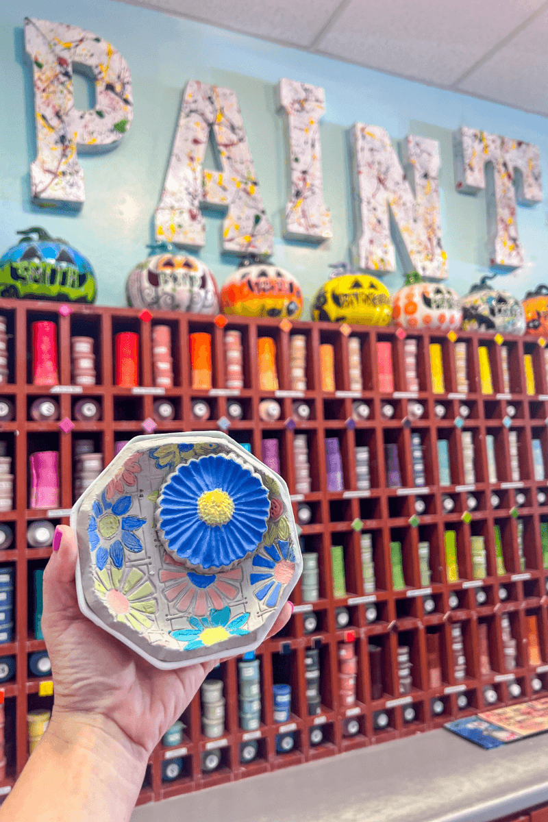 Create, Connect & Get a Little Messy at This Memphis Pottery Studio - 3 A hand holds a painted ceramic dish with a blue flower in front of a paint display wall with colorful paint bottles and decorated letters spelling “PAINT.”