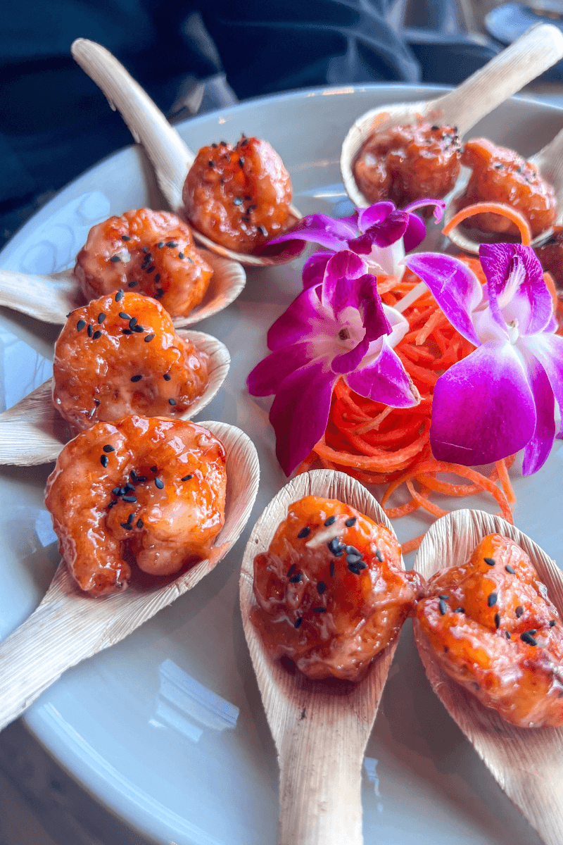 Did You Know You Could Spend the Night in the Memphis Pyramid? - 4 Six shrimp with sauce and black sesame seeds are served on wooden spoons, garnished with shredded carrots and two purple orchids on a white plate.