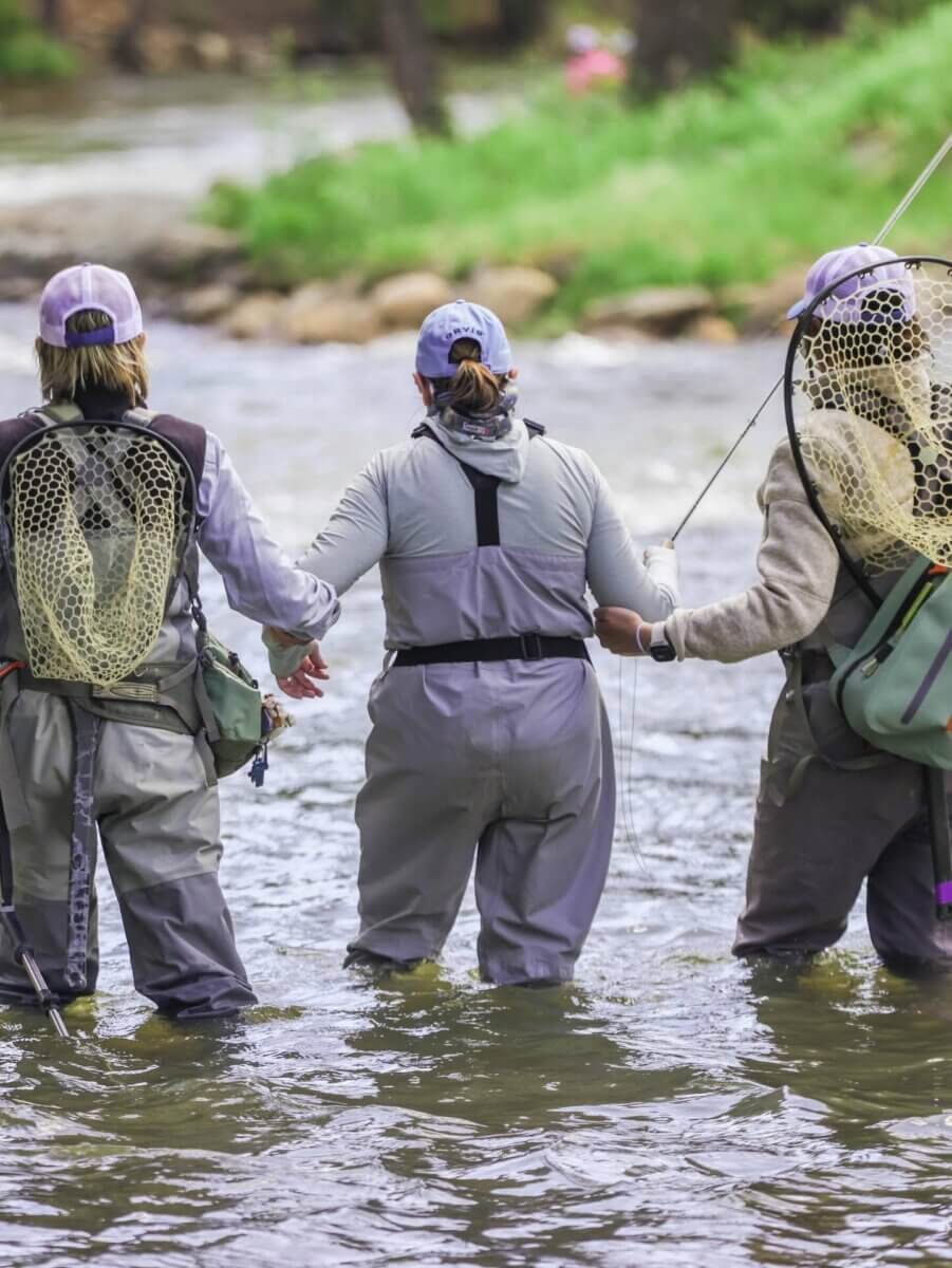 Breast Cancer Sucks. 6 Surprising Ways to Support Survivors - 3 Three people in fishing gear stand in a shallow river, holding hands and facing away from the camera—an inspiring moment that captures the strength and unity of breast cancer survivors.