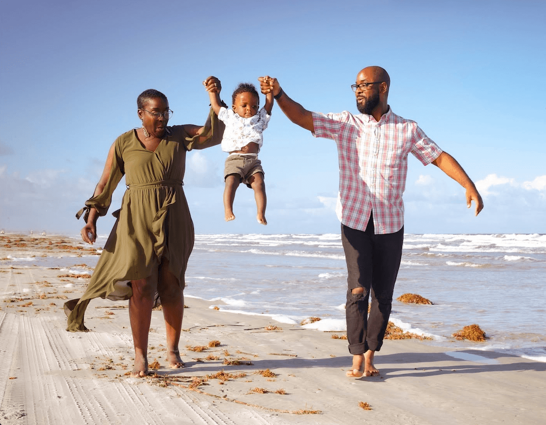 Breast Cancer Sucks. 6 Surprising Ways to Support Survivors - 2 A woman and man walk barefoot on a beach, swinging a smiling toddler between them. The sky is clear and waves are visible in the background, capturing a joyful moment shared by breast cancer survivors embracing life with their family.